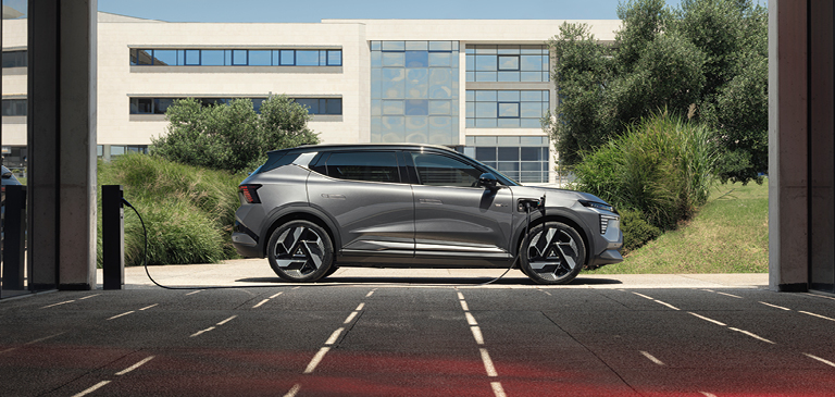 Grauer Mitsubishi Eclipse Cross an einer Ladesäule, seitlich im Profil zu sehen, während er vor einem modernen Gebäude mit viel Glas und Grünflächen im Hintergrund lädt.